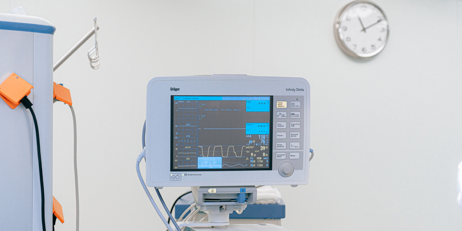 A heart monitor that is plugged into another, cylindrical piece of machinery. It is a pale grey colour and is against a grey wall. A clock hangs in the top right.