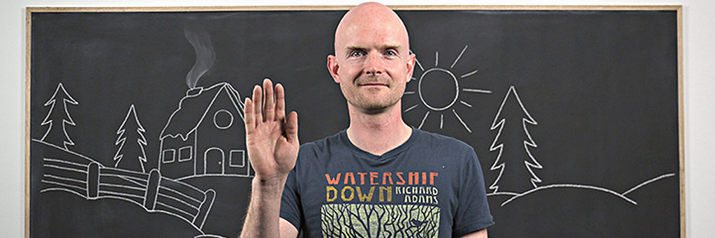 Dr. Steven Barnes standing in front of a stylized image of a chalkboard, with chalk drawing of a cabin and trees in the woods. He is white and is smiling and holding up one hand in a greeting. He is wearing 
a Watership Down t-shirt.