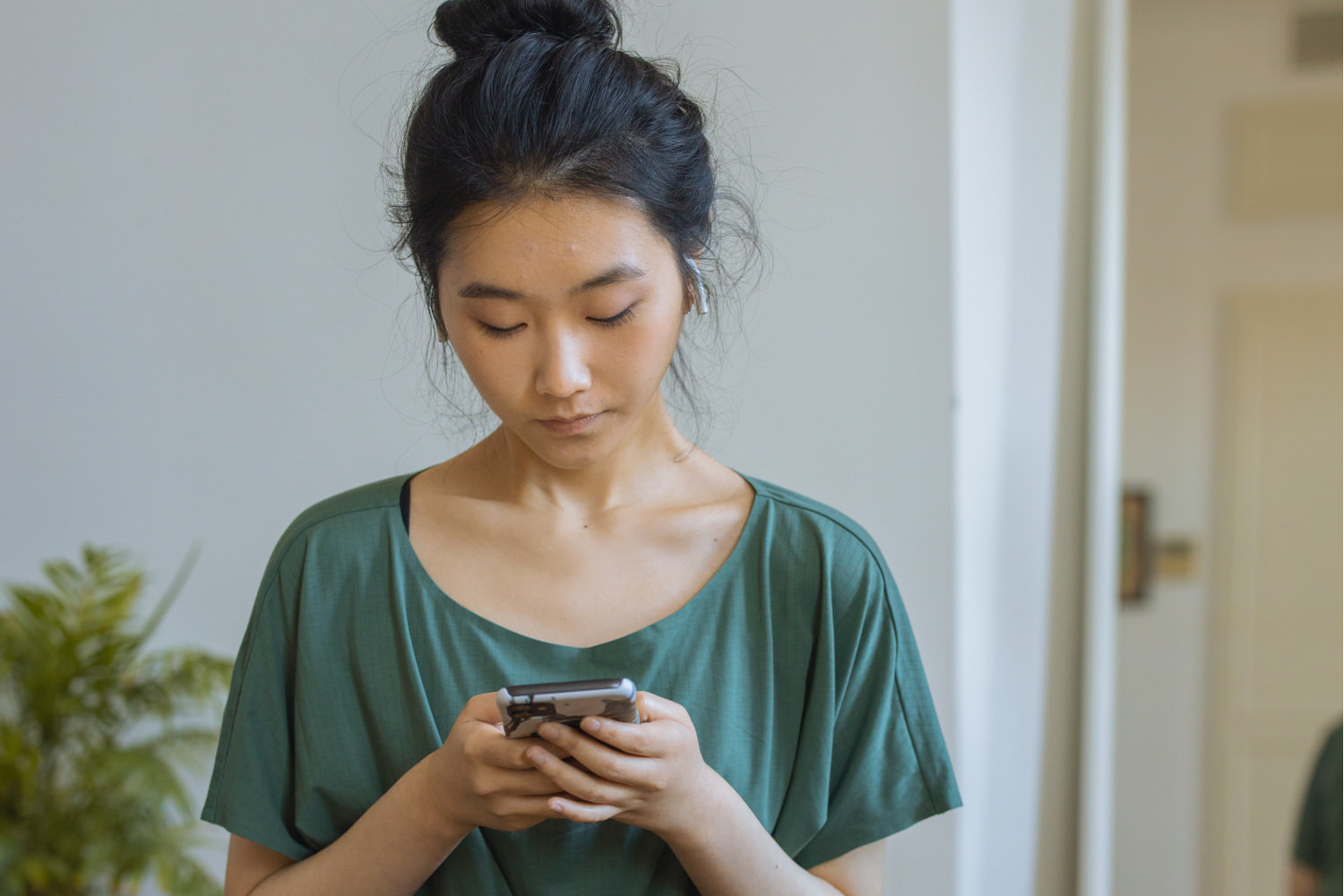 A young east asian woman is holding her phone and looking down at it. She has a messy bun and is wearing a loose-fitting green t-shirt. She looks calm and like she might be selecting a meditation.