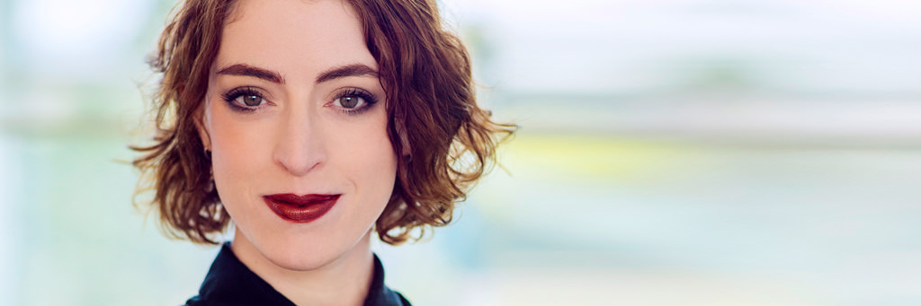 A headshot of Emma smiling against a blurred greenish background, like a window out of focus. She is Caucasian and has chin-length wavy red hair.