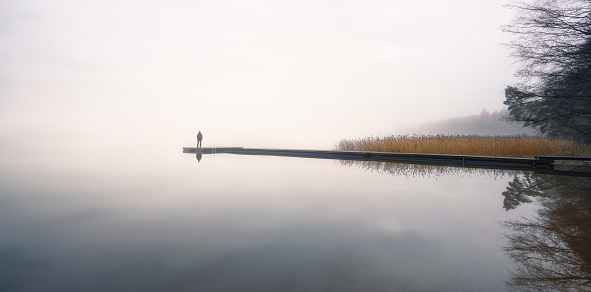 A solitary individual stands at the very edge of a narrow dock. It is on a grey lake and extends from an island. The picture is zoomed out and the figure is tiny. The sky is cloudy.