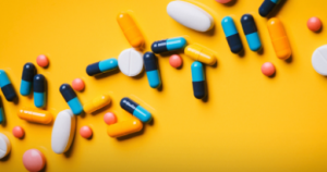 Image has a bright yellow background. Medication pills and tablets run across the photo from the bottom left to top right. The medication is pink, yellow, white and blue. 