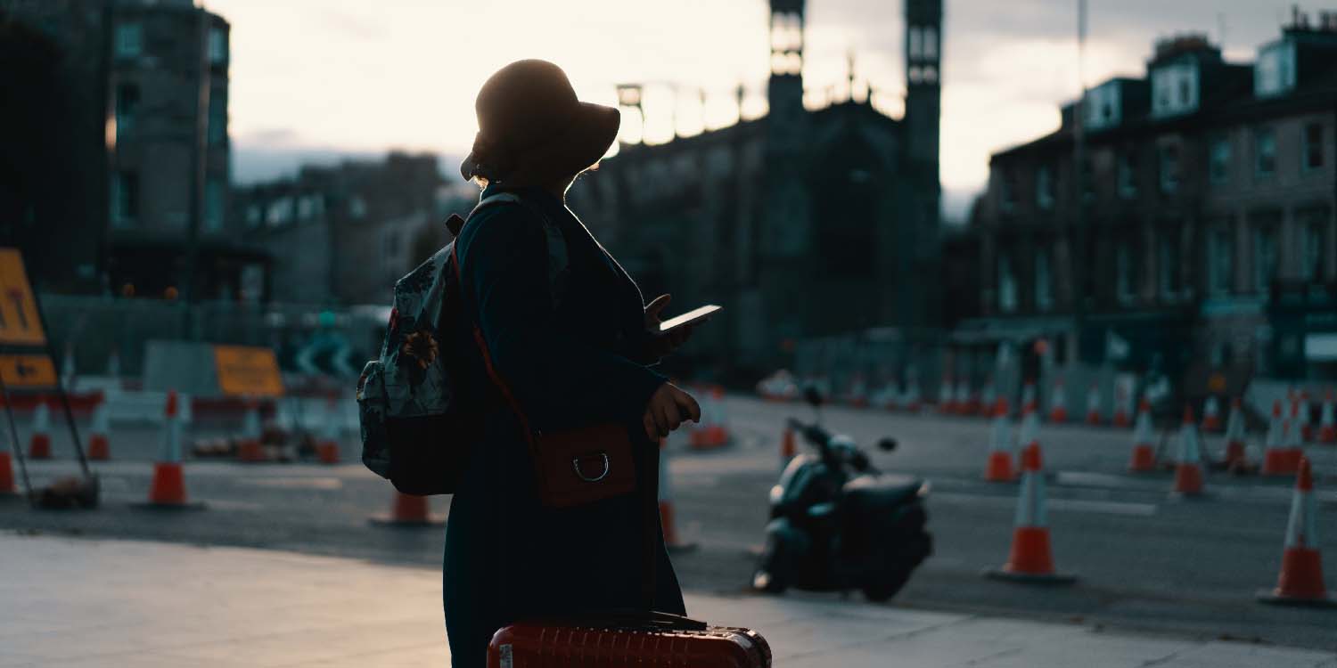 A woman is shown from the back holding a red suitcase. She is wearing a long coat, a backpack with a flower pattern, and an orange hat with a small floppy brim. She is holding her phone. In front of her, there are European buildings and many, many traffic cones. It looks like a beautiful city, but there is a lot that is being worked on.