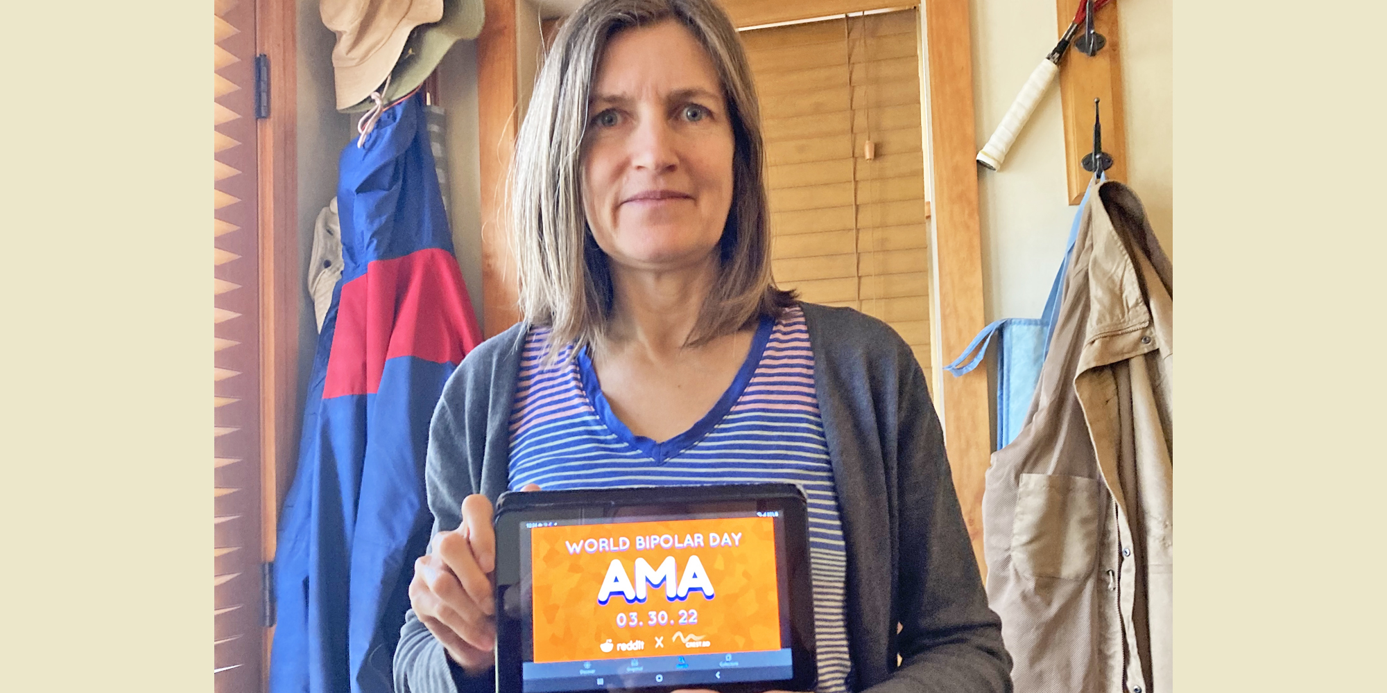 Keri is a room with cream walls and a light brown woode frames window with matching curtains. She is wearing a horiztonal striped blue and red T-Shirt and a grey cardigan. She is Caucasian and has shoulder length light brown hair. In her hands is a tablet displaying the AMA poster.