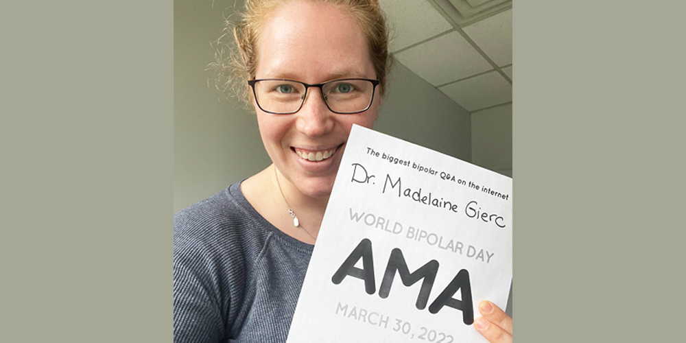Madelaine is taking a selfie. She is Caucasian and has her red hair pulled back. She is wearing a silver chain with one white pearl, and a blue sweater. In her left hand she is holding the AMA sign.