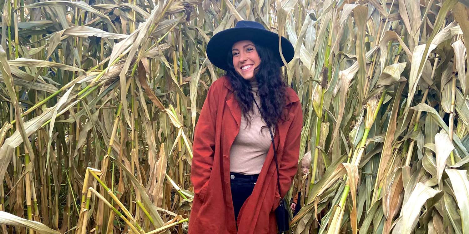 Naomi is standing in a field of corn. She has light skin and is wearing a wide-brimmed black hat, a long red jacket, a pale pink top, and black jeans. Her hair is long and dark.