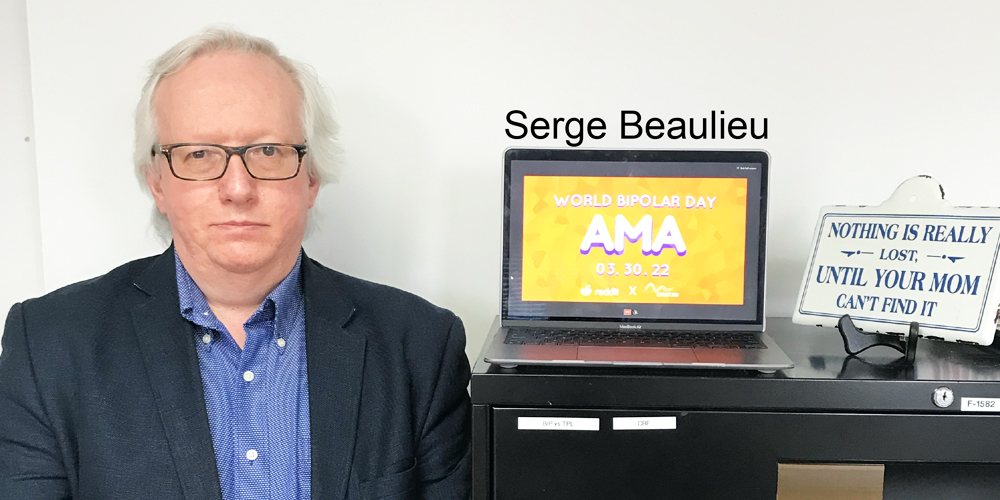 Serge is standing to the left of a black filing cabinet. He is wearing a blue and white dotted button up shirt with a black suit jacket. He has white hair and rectangular glasses. On the filing cabinet is a laptop displaying the AMA poster and a sign that reads "nothing is really lost, until your mom can't find it."