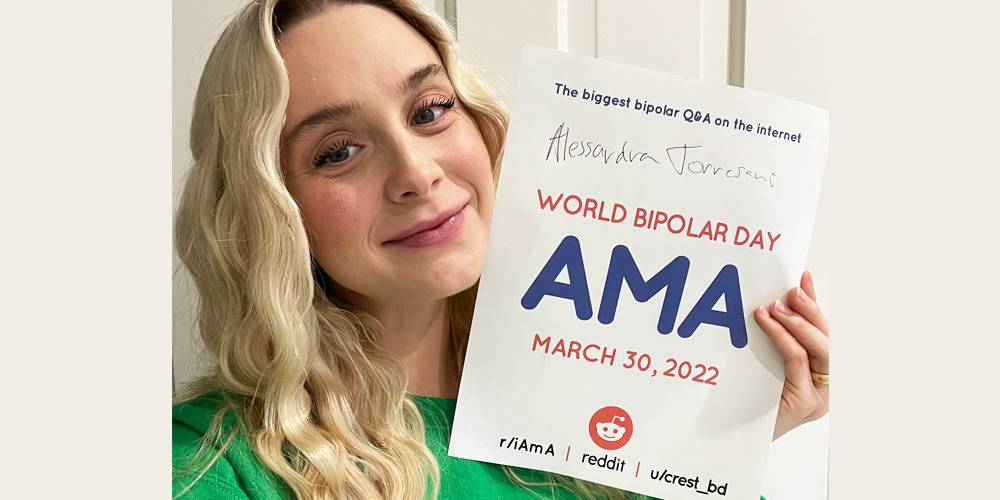 Alessandra is standing indoors in front of a white door. Alessandra is Caucasian and has long platinum blonde hair set in waves. She is wearing a green sweater, and is holding an AMA proof sign. 