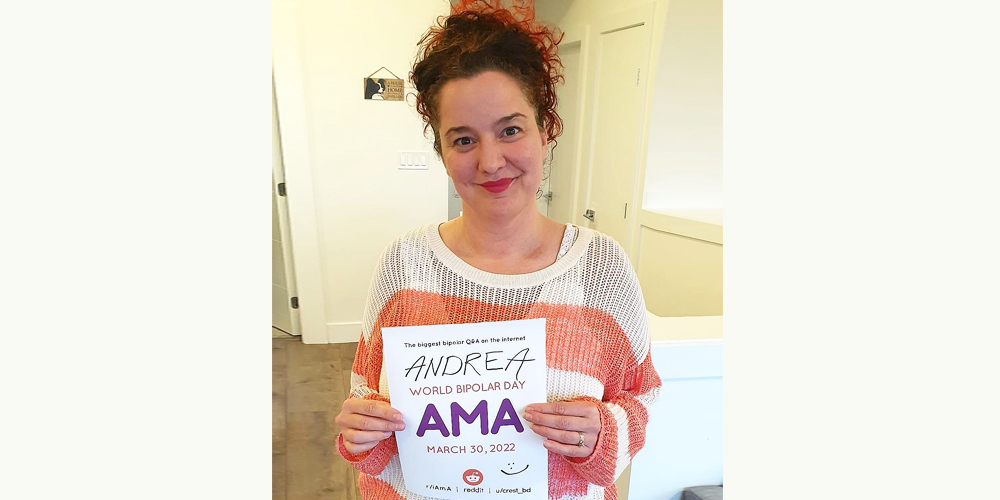 Andrea is in a room with beige tiles and pale yellow walls. She is a white woman with red hair wearing a red and white striped long sleeve shirt. In her hands she is holding the AMA sign.