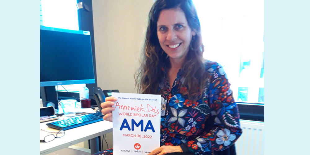 Annemiek is sitting indoors in an office. The her left is her desk with her computer and glasses The is a window behind her which appears to look out to another building. Annemiek is Caucasian and has long brown hair. She is wearing a zip-up jacket with vibrant blue, red, and white flowers, and is holding an AMA proof sign. 