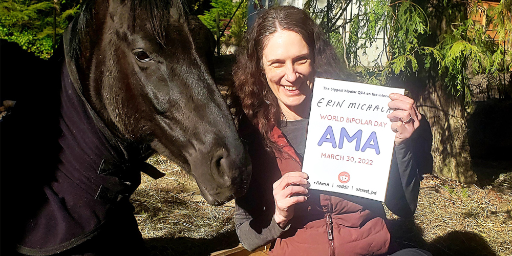 Erin outdoors with two cedar trees in the background. She is Caucasian and has curly dark brown hair. On the left of the photo is her horse, Vale, who is a glossy black colour. Erin is wearing a black shirt with a dark red vest. She is smiling warmly and holding a sign proving she will be involved in the bipolar ama, with her name and the AMA date and time on it.
