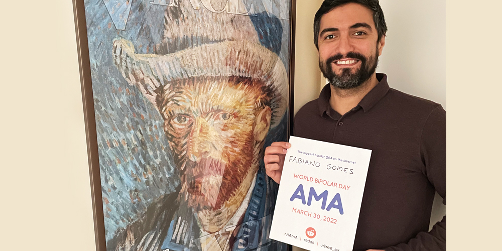 Fabiano is standing indoors in front of a self-portrait of Vincent Van Gough. He has olive-toned skin and has black hair and a close-shaved black beard. He is wearing a brown dress shirt and is holding an AMA proof sign.