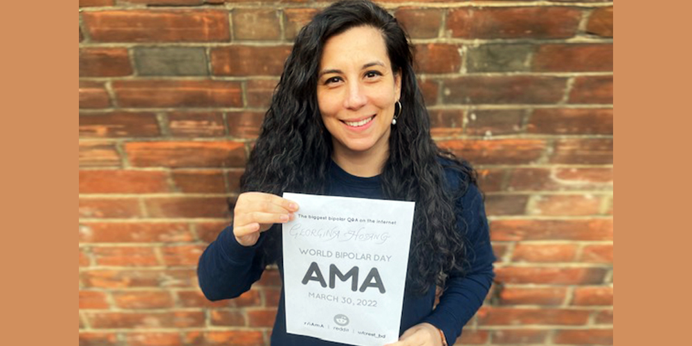 Georgina is standing in front of a red brick wall. She has long dark curly hair and is wearing a long sleeve blue shirt. In her hands, she is holding the AMA sign.