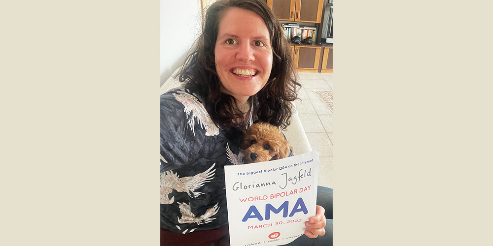 Background is a white wall and white tiles. Glorianna is wearing a blue shirt with white birds and is sitting on a white couch. In her lap is a cute little dog. in her left hand she is holding the AMA poster.