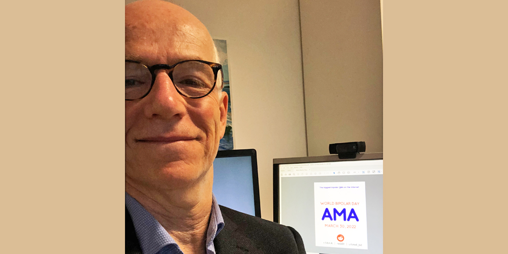 Greg is standing infront of a desktop monitor with the AMA sign on it. He is wearing a black suit jacket and a light blue collared shirt. He is bald and has round brown glasses on.