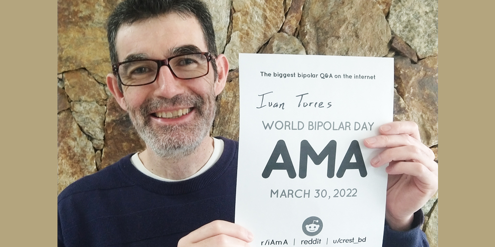Ivan is in front of a stone wall. He has short brown hair, a salt and pepper beard and is Caucasian. He is wearing square brown glasses and white T-Shirt under a blue sweater. He is holding to his left the AMA sign.