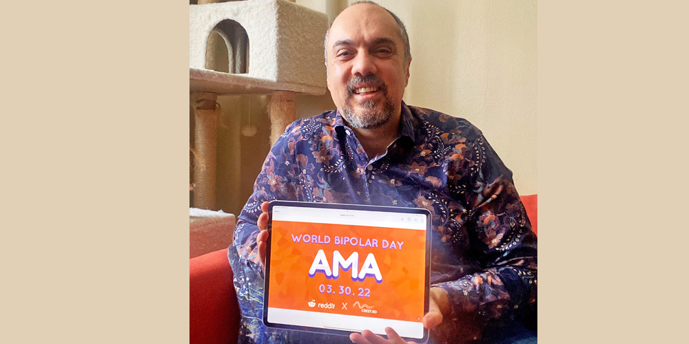 Kamyar is sitting in a room with white walls sitting on a red chair. He is bald with a salt and pepper beard. He is wearing a colourful, patterened, red and purple collared shirt. In his hands, he is holding an iPad with the AMA sign on it's screen.