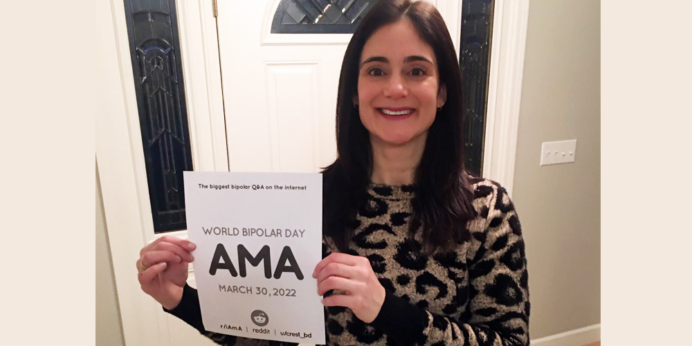 Lisa is in a hallway with a white door behind her. She is wearing a cow print long sleeve shirt and has long brown hair. She is smiling and holding the AMA sign to her right side.