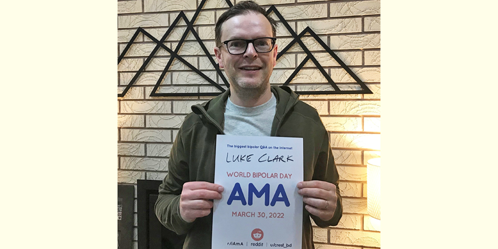Luke is in a room with cream brick walls. He is a white man with round glasses, wearing a grey T-shirt and dark green hoodie. His hair is short and brunette. He is holding the AMA sign. 