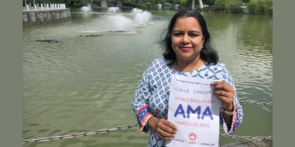 Nigila is standing in front of a lake with a water fountain in it. She is wearing a light blue patterned shirt with red and blue cuffs. She has shoulder-length black hair and is holding the AMA sign with both hands.