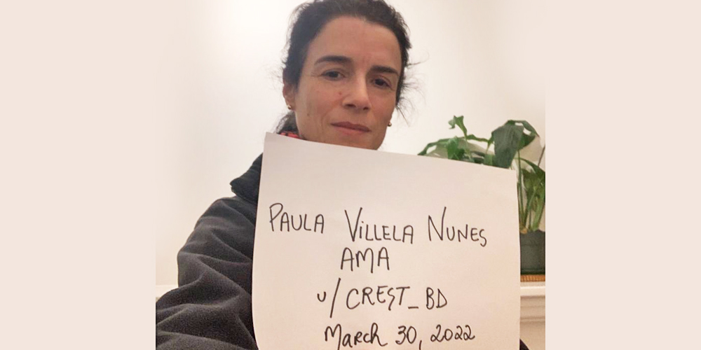Paula is standing indoors holding a proof sign. There is a houseplant behind her. She has olive-toned skin and long, dark hair pulled back in a pony tail.