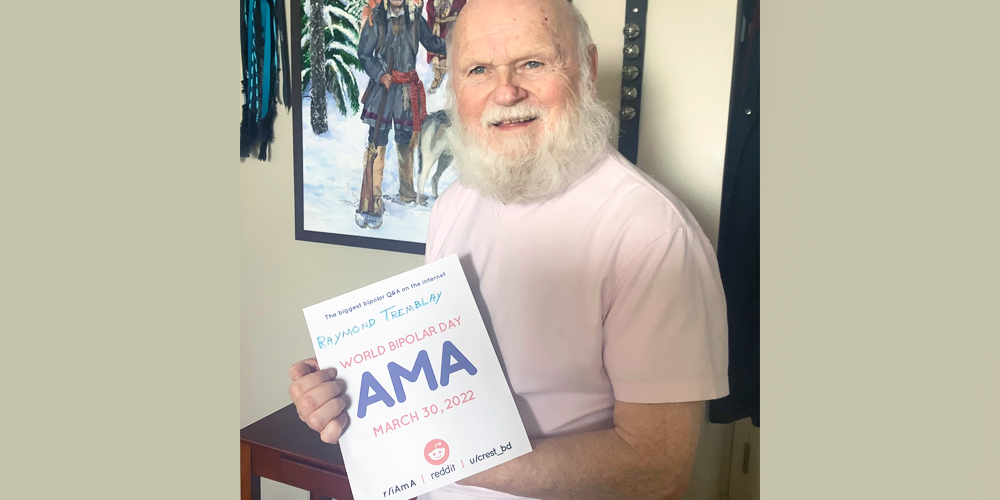 Raymond is sitting indoors on a wooden bench. There is a picture of kids in the snow behind him. He is Caucasian and has a big white beard. He is wearing a white t-shirt and holding an AMA proof sign.