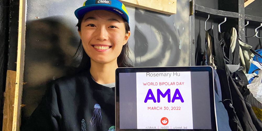 Rosemary is standing in an industrial-looking building with coat closet and a sheet of plastic behind her. She is Chinese Canadian and is holding a laptop with the AMA proof and her typed name up on the screen. She has shoulder-length, black hair, and is wearing a teal and yellow ballcap and a black sweater with a jellyfish pattern.