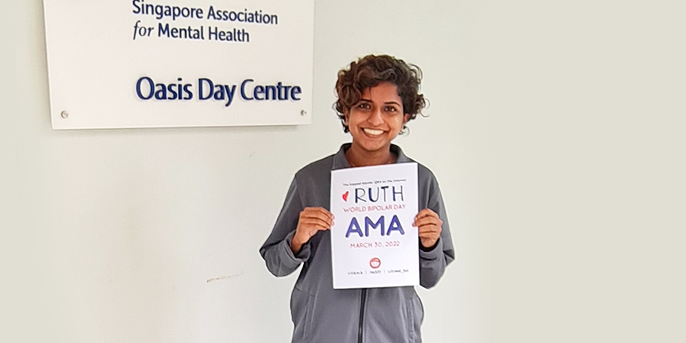 Ruth is in a room with a sign for the Singapor Association for Mental Health Oasis Day Centre and grey walls. She is wearing a grey button up shirt and has short black curly hair. She is holding the AMA sign in her hands.