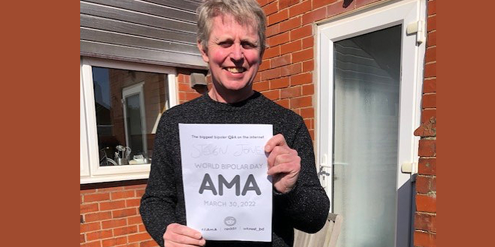 Steve is standing outside of a red brick house with a white framed glass door. He is wearing a black shirt and hold a World Bipolar Day AMA sign.