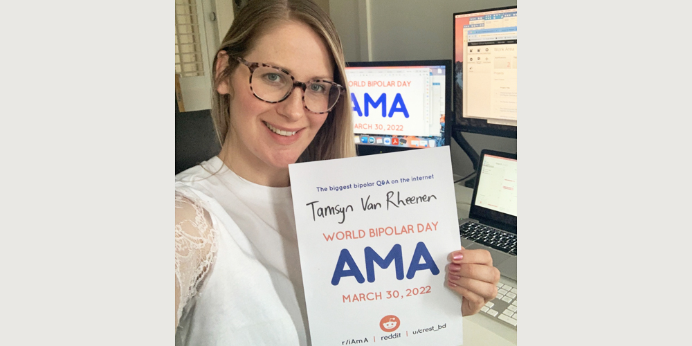 Tmasyn is in an office with a monitor and computer on a white desk. She is a white woman wearing a white high collared shirt with sheer white sleeves. She has blonde, long hair and square brown glasses. In her left hand she is holding the AMA poster.