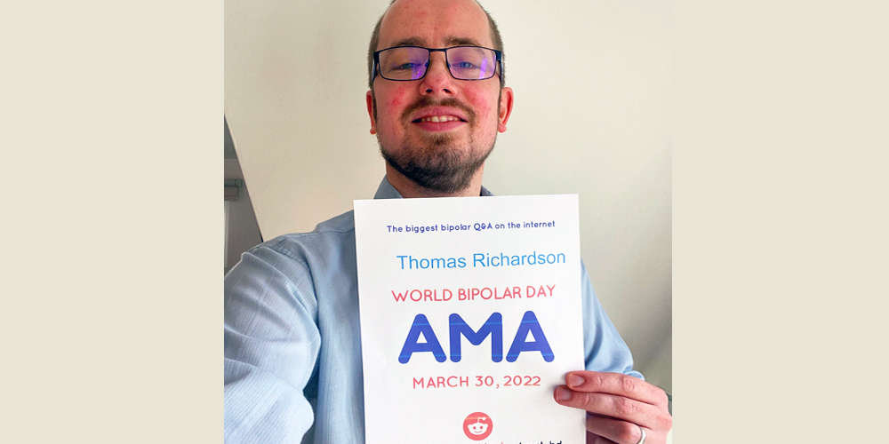 Thomas is standing in front of a beige walling holding up the AMA proof sign. He is Caucasian, with short, close-cropped brown hair and a slight beard. He is wearing a pale blue dress shirt and black-framed, rectangular glasses.