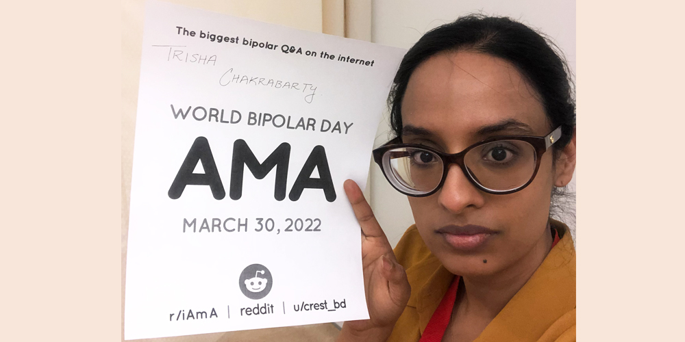 Trisha is in a room with white walls. She has black hair and brown oval glasses. she is wearing a yellow and red v neck shirt and holding the AMA sign in her right hand.