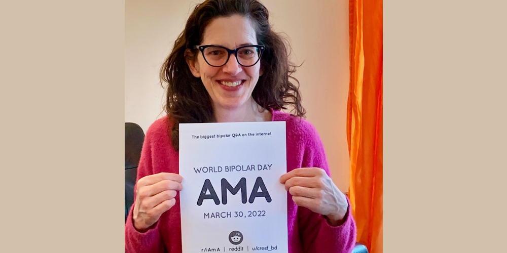 Victoria is standing in a room with white walls and a window covered by orange curatins. She has curly, shoulder length brown hair and is wearing round brown glasses and a long sleeve pink shirt. In her hands she is holding the AMA sign.