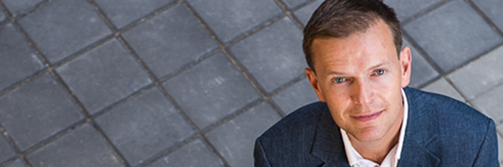 Luke is a white man with short, light brown hair. He is looking up towards the camera, and is wearing a dark blue suit jacket with a white button up shirt. Only his shoulders and head are visible. The background is a grey tile floor. 