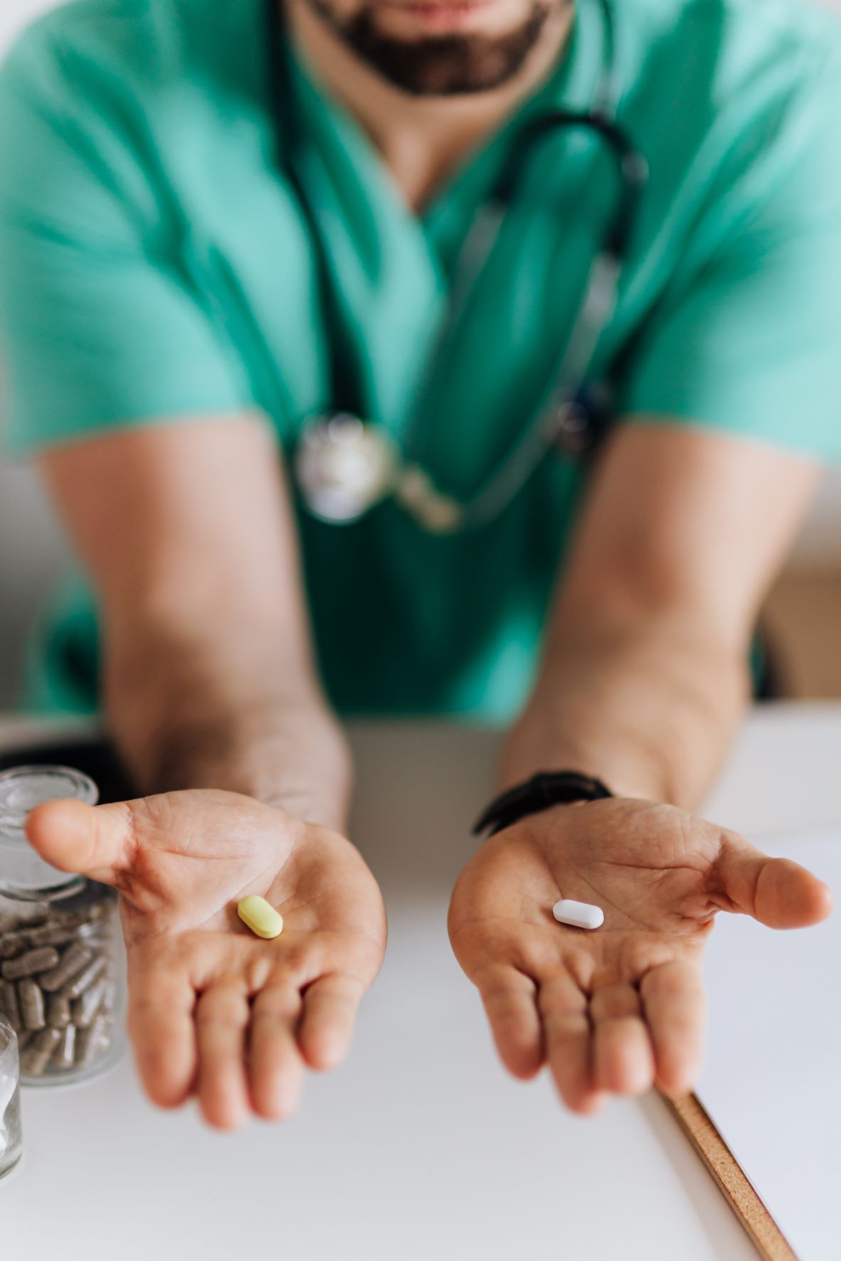 In the foreground of the photo are a man's hands stretched out towards the viewer. In the left is a small yellow pill, and in the right is a small white pill. In the background the man is wearing turquois scrubs and a stethoscope around his neck. Only his chin is visible, which has a dark brown beard. 