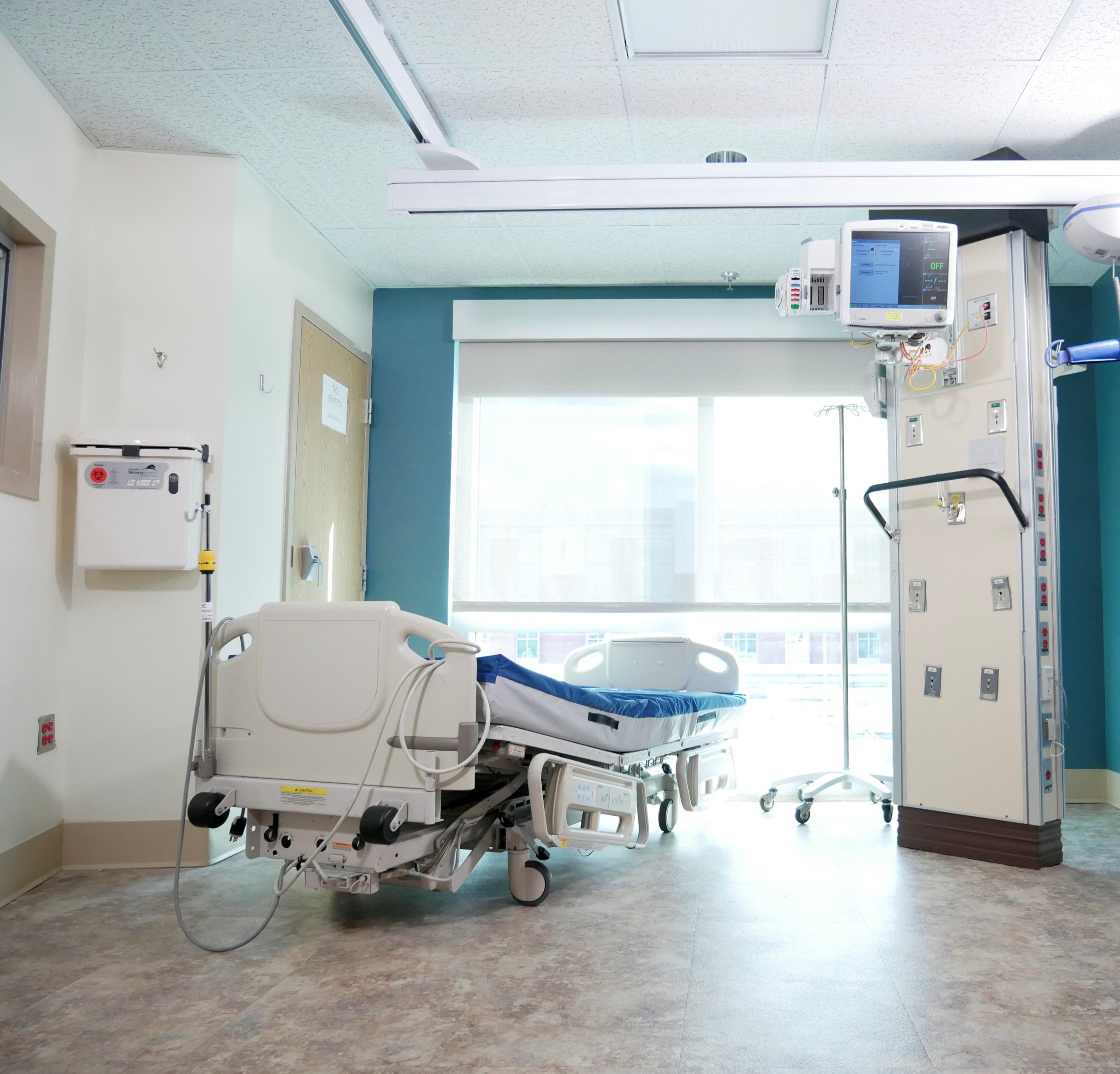 Image is of a hospital room with white walls and a large window. Facing the windo is a blue, adjustable hospital bed with blue sheets. Next the to the bed is a small T, hanging on a white pillar in front of the window.