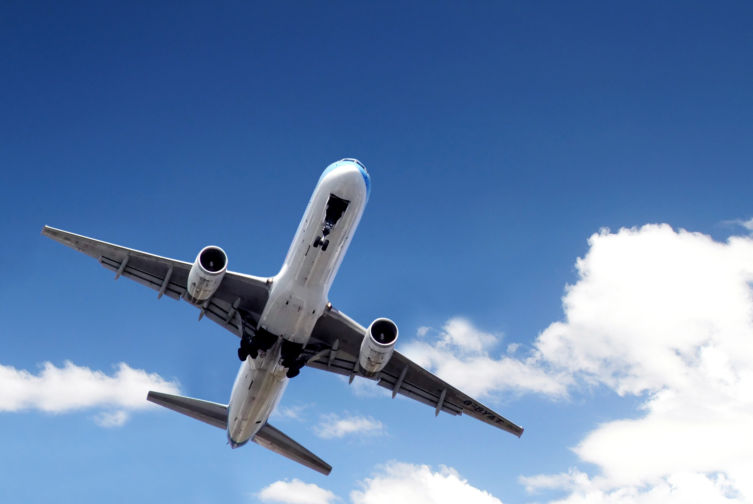 Image foreground is of a plane taking off, the view is from below. The background is a blue sky with a few clouds.