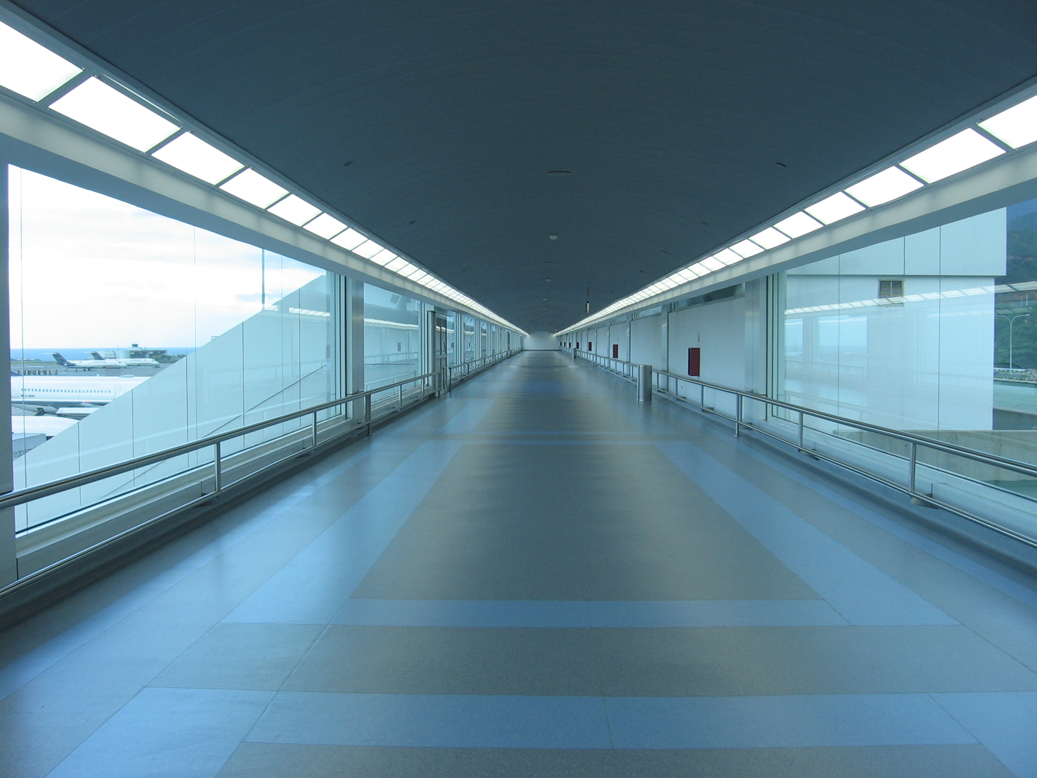 Empty airport hallway, lined with windows. The ground is grey with thick blue lines. 