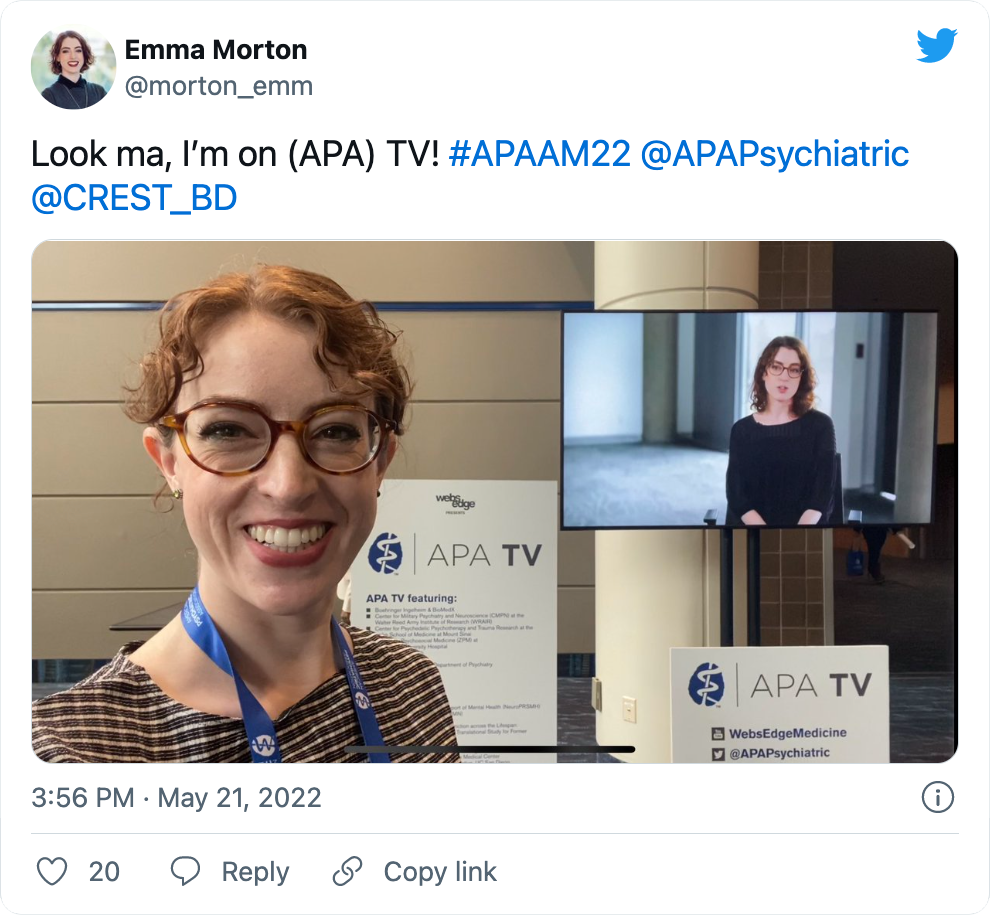A tweet from Dr. Emma Morton (emm_morton on Twitter) that says 'Look Ma, I'm on (APA) TV!' Attached is a picture of Emma standing in front of a flat-screen TV playing a video of her. She is Caucasian with shoulder-length, wavy red hair and is wearing glasses. On the TV, she is wearing a black shirt and appears to be explaining something. The 'real' Emma is smiling and wearing a tan shirt and a lanyard. It says the Tweet was posted May 21, 2022, and it has 20 likes.