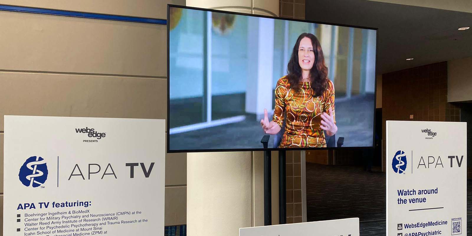 A flatscreen TV is hung on a pillar in what looks like a hotel lobby. On the screen, Erin is in the middle of talking. She is Caucasian with long, wavy brown hair and is wearing a shiny brown, orange and green desk. There are signs to the left and right of the TV that say 'APA TV.'