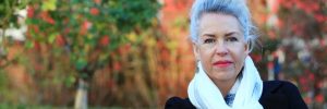 Blurred background of trees in the fall. Felice Jacka is in the foreground. She is a white woman wearing a white scarf and short grey hair.
